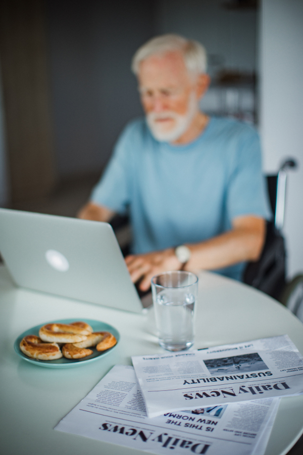 Senior man in a wheelchair shopping online on laptop. Elderly man using digital technologies, working on a notebook in his kitchen. Concept of seniors and digital skills.