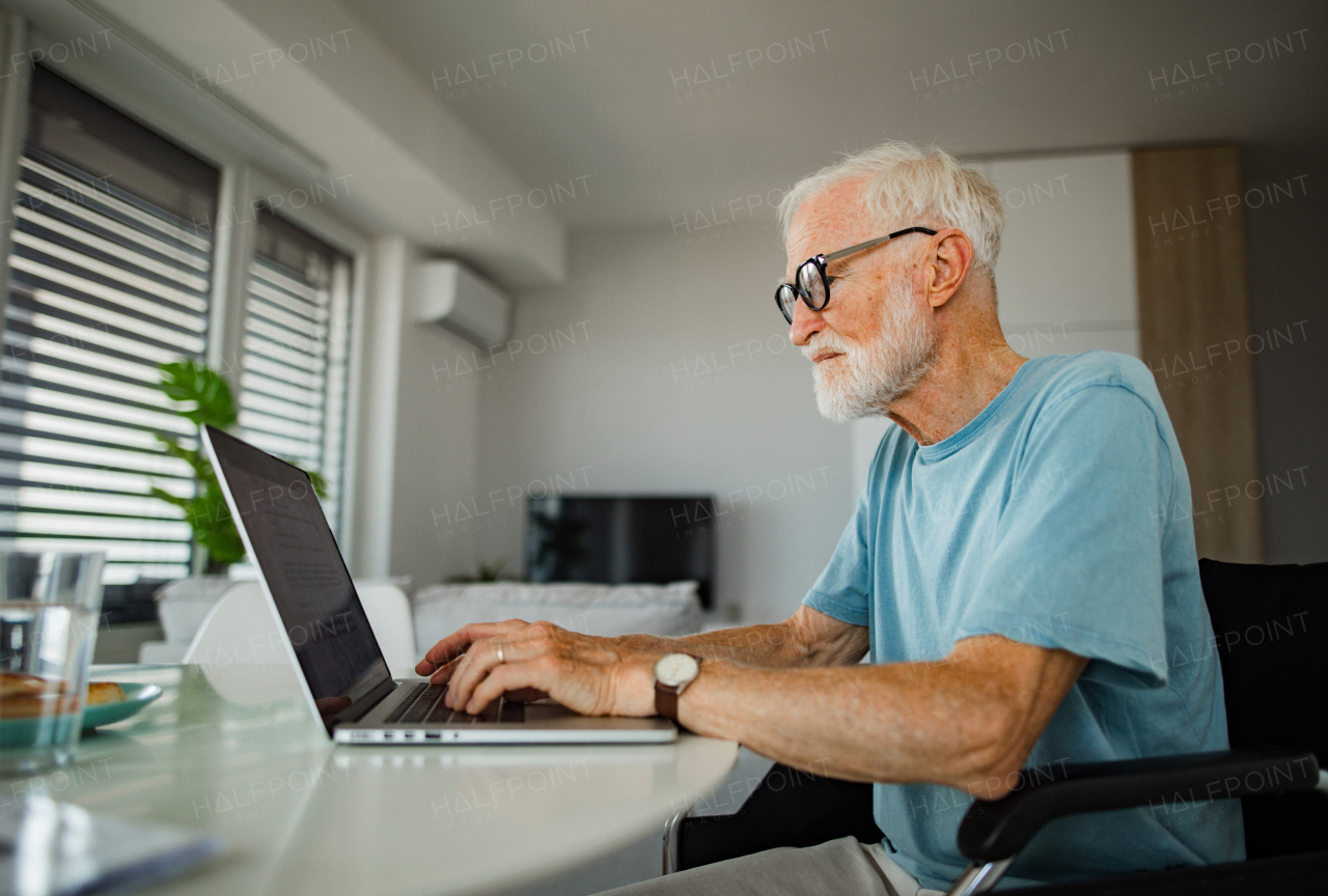 Senior man in a wheelchair working from home during retirement. Elderly man using digital technologies, working on a laptop. Concept of seniors and digital skills.