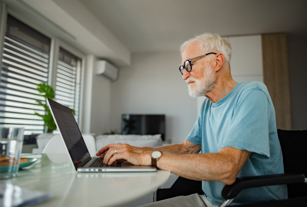 Senior man in a wheelchair working from home during retirement. Elderly man using digital technologies, working on a laptop. Concept of seniors and digital skills.