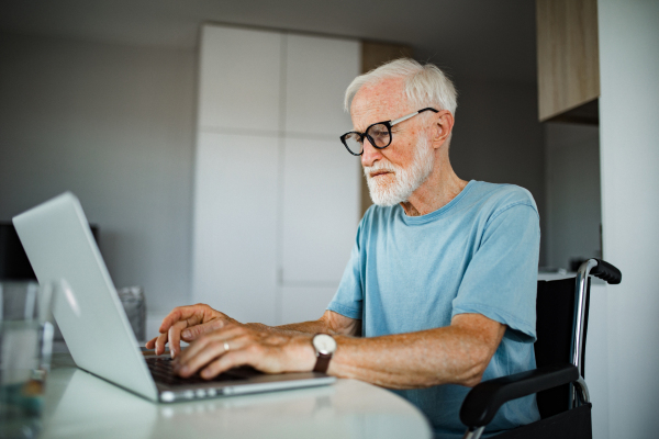 Senior man in a wheelchair working from home during retirement. Elderly man using digital technologies, working on a laptop. Concept of seniors and digital skills.