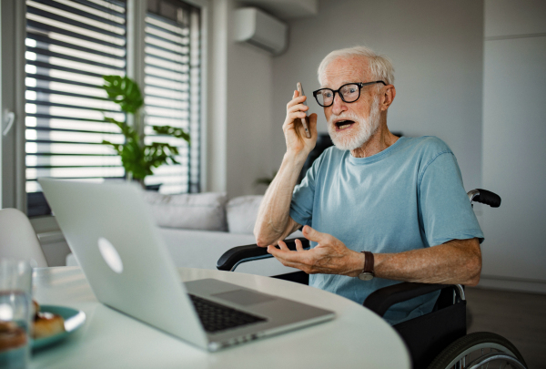 Senior man in a wheelchair working from home during retirement. Elderly man using digital technologies, working on a laptop and making phone call at home. Concept of seniors and digital skills. Senior calling customer service to complain.