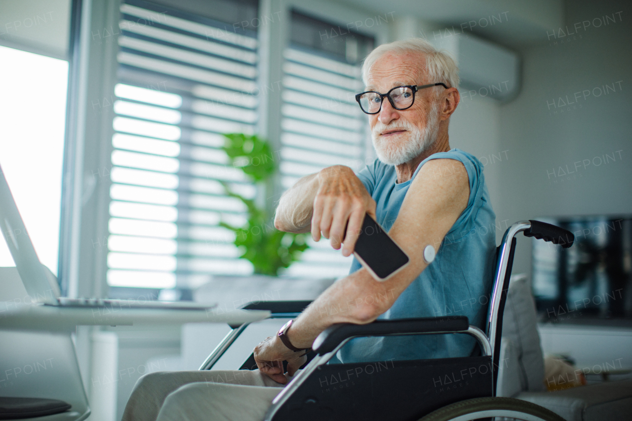 Diabetic senior man checking blood glucose level at home using continuous glucose monitor. Elderly man connecting his CGM with smarphone to see blood sugar levels in real time.