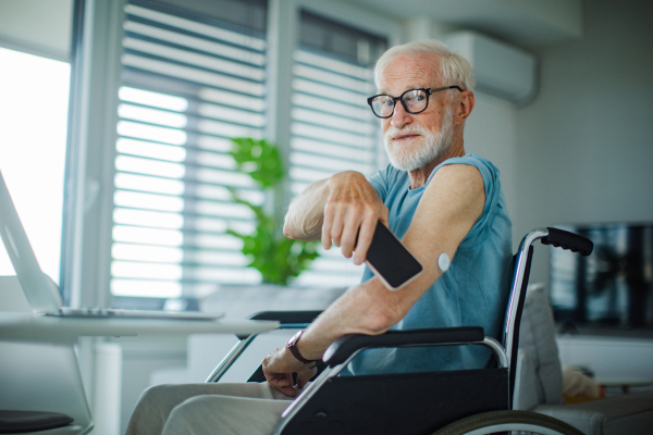 Diabetic senior man checking blood glucose level at home using continuous glucose monitor. Elderly man connecting his CGM with smarphone to see blood sugar levels in real time.