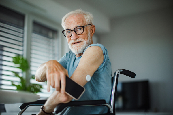 Diabetic senior man checking blood glucose level at home using continuous glucose monitor. Elderly man connecting his CGM with smarphone to see blood sugar levels in real time.