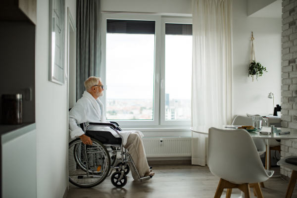 Senior man in a wheelchair spends time alone in his apartment. Concept of loneliness and dependence of retired people.