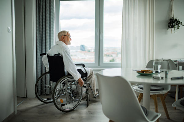 Senior man in a wheelchair spending time alone in his apartment. Concept of loneliness and dependence of retired people.