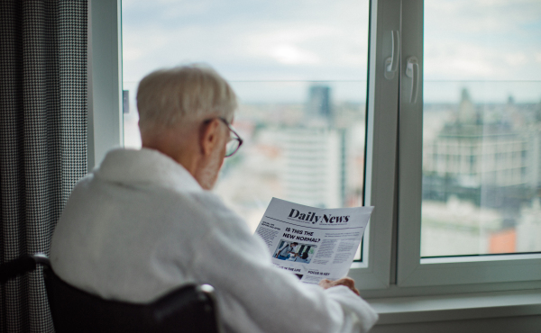 Rear view of elderly man in a wheelchair reading the newspaper in his robe in the morning. Concept of loneliness and dependence of retired people.