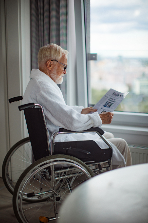 Elderly man in a wheelchair reading the newspaper in his robe in the morning. Concept of loneliness and dependence of retired people.