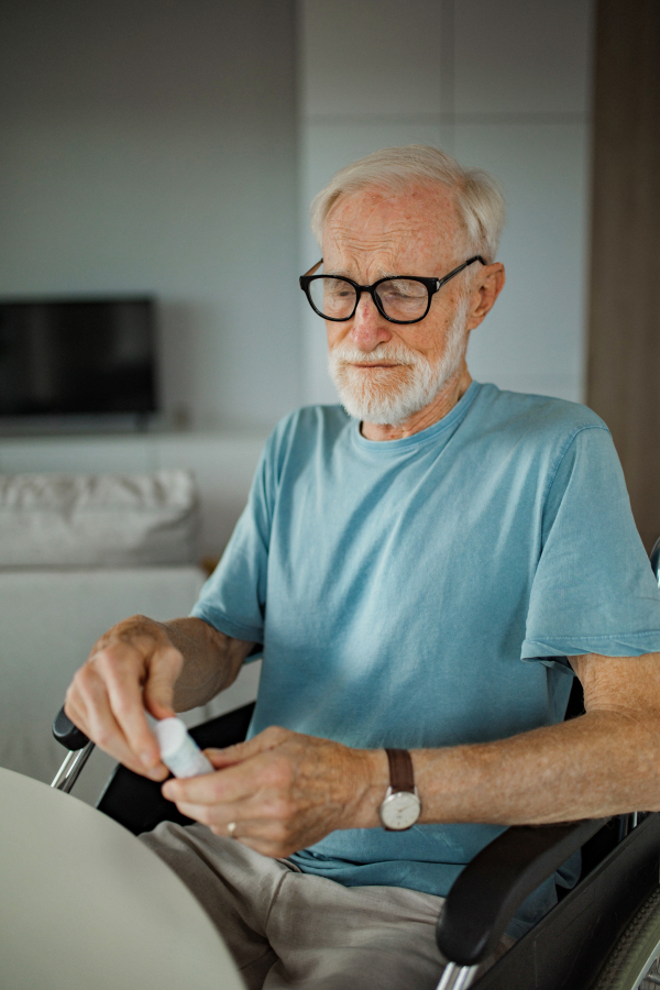 Senior man in wheelchair taking pills at home. Chronically ill man taking medication, reading the label on the bottle.