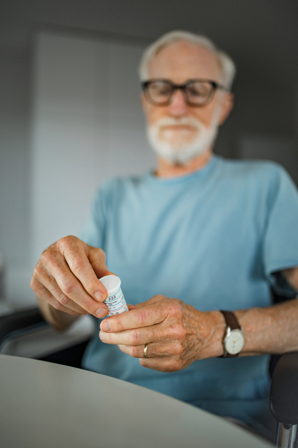 Senior man in wheelchair taking pills at home. Chronically ill man taking medication, reading the label on the bottle.