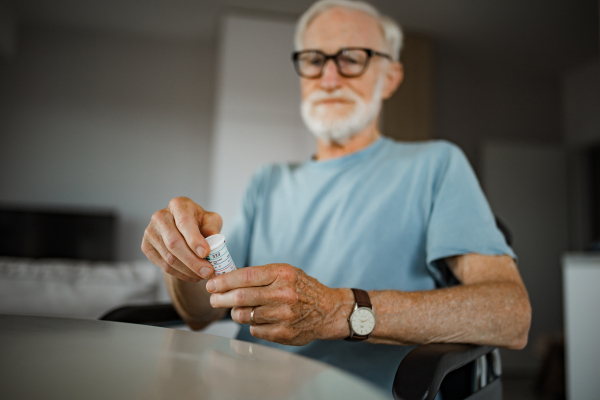 Senior man in wheelchair taking pills at home. Chronically ill man taking medication, reading the label on the bottle.