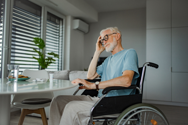 Portrait of senior man in wheelchair taking pills at home, he is confused about mediacation usage. Chronically ill man have no money for his medication.