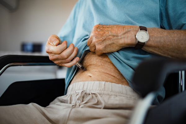 Diabetic senior patient injecting insulin in his belly. Close up of senior man in wheelchair with type 1 diabetes taking insuling with syringe needle.