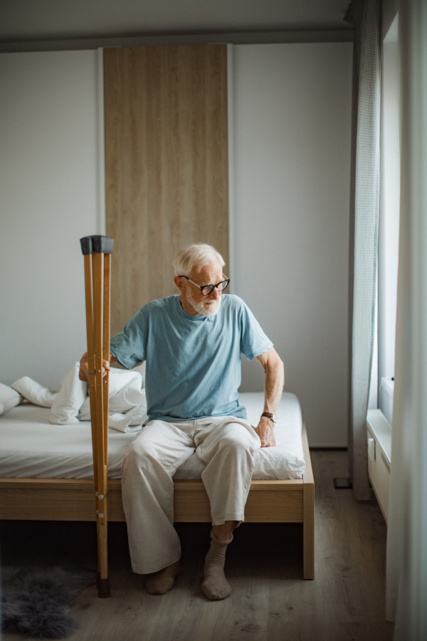 Portrait of an elderly man with crutches trying to stand up from bed.