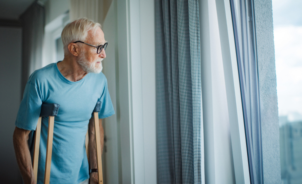 Sad senior man with crutches spending time alone in his apartment, looking out of window. Concept of loneliness and dependence of retired people.