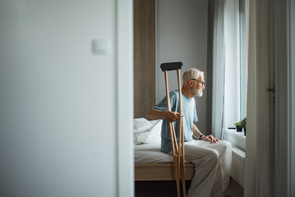 Sad senior man with crutches spending time alone in his apartment, looking out of window. Concept of loneliness and dependence of retired people.