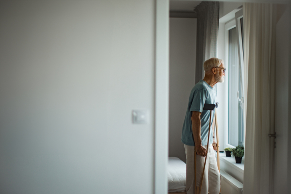 Sad senior man with crutches spending time alone in his apartment, looking out of window. Concept of loneliness and dependence of retired people.