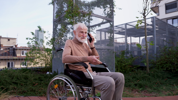 Portrait of senior man in a wheelchair sitting outside in an urban garden, making call with smartphone. Video of a elegant elderly man with gray hair and beard in rooftop garden in the city at autumn.