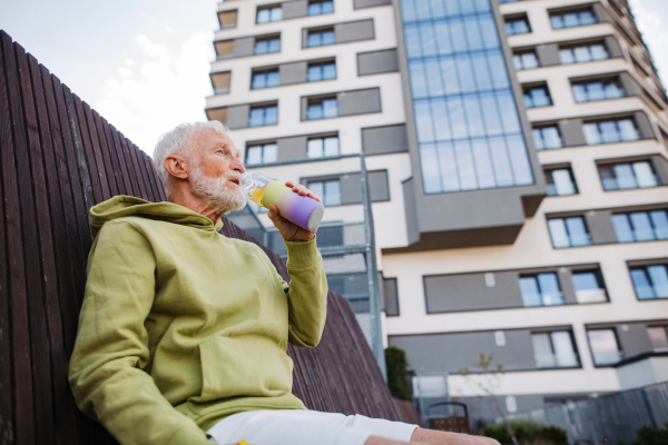 Senior man exercising outdoors in the city. Elderly man sitting on bench, drinking water after his morning run. Low angle shot.