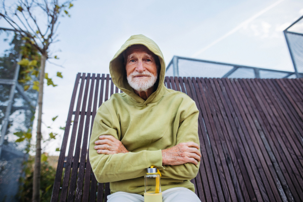 Portrait of cool elderly man in a hoodie with the hood on his head and arms crossed over his chest, during exercising outdoors.