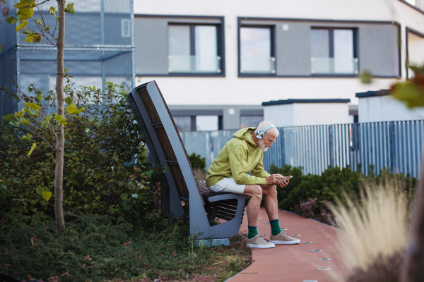 Senior man exercising outdoors in the city. Elderly man resting on a bench, listening to music through wireless headphones, preparing for his morning run.