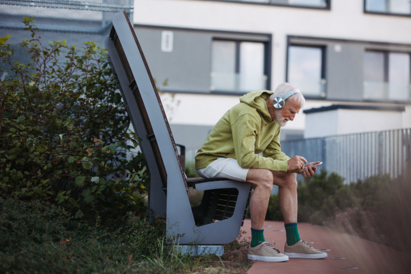 Senior man exercising outdoors in the city. Elderly man resting on a bench, listening to music through wireless headphones, preparing for his morning run.