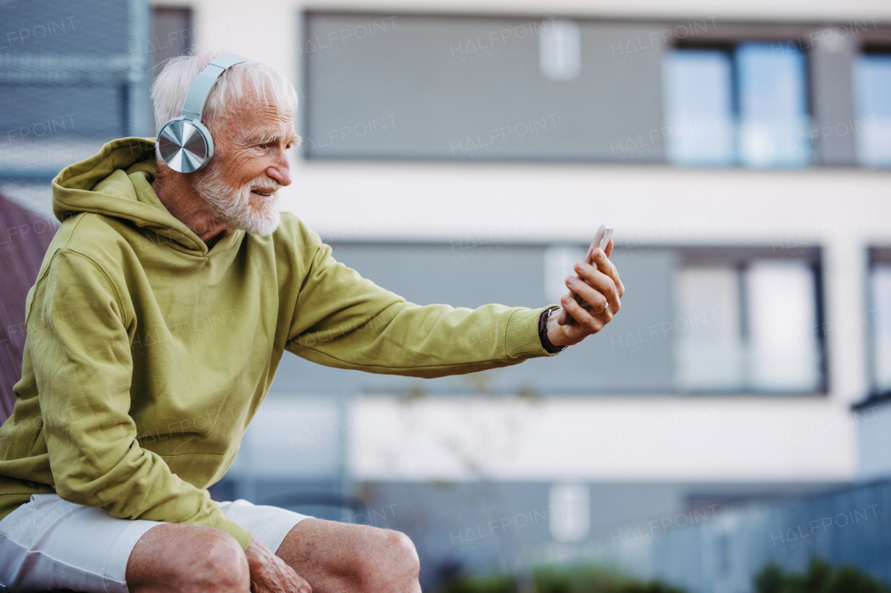 Senior man exercising outdoors in the city. Elderly man resting on a bench, listening to music through wireless headphones, taking selfie.