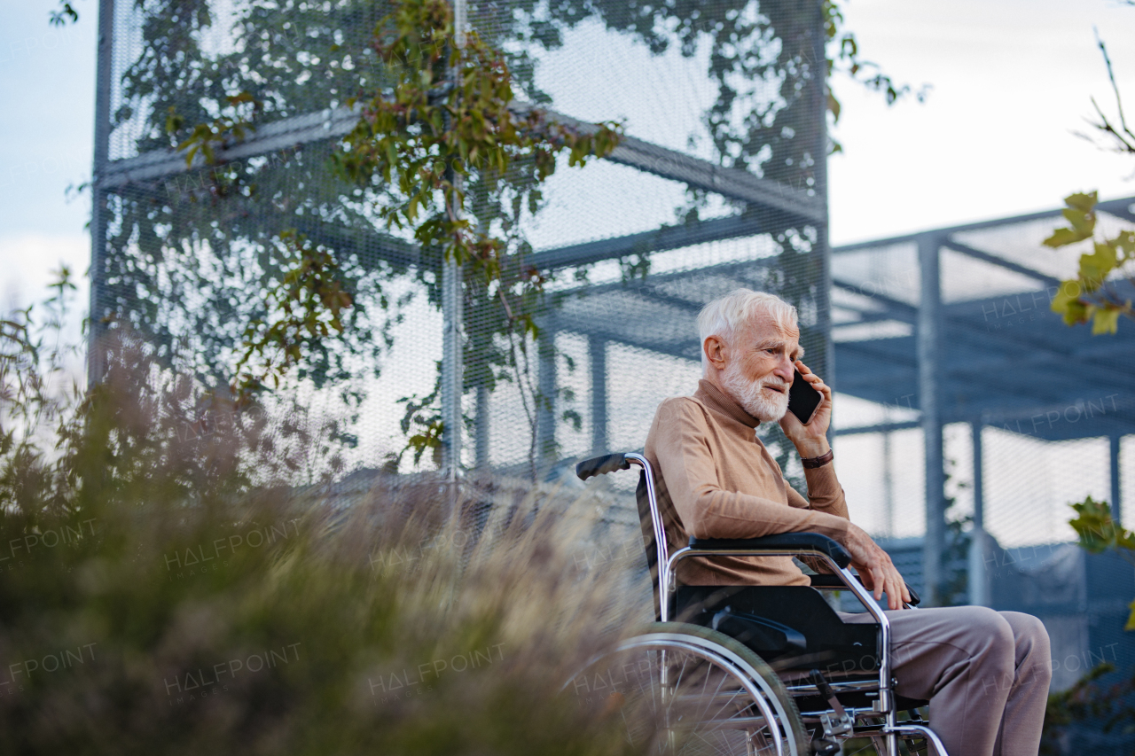 Portrait of senior man in a wheelchair sitting outside in an urban garden, making call with smartphone. Portrait of a elegant elderly man with gray hair and beard in rooftop garden in the city at autumn.