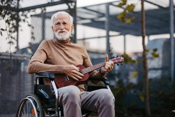 Senior man sitting in a wheelchair outdoors, playing the ukulele. Retired musician playing the guitar, remebering about old times. Therapeutic effects of music on elderly people.