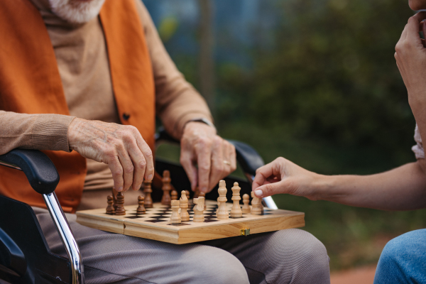 Close up of senior man playing chess outdoors with his daughter. Nursing home client in wheelchair spending quality time with caregiver, enjoying in a game of chess and conversation.