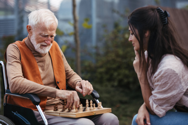 Senior man playing chess outdoors with his daughter. Nursing home client in wheelchair spending quality time with caregiver, enjoying in a game of chess and conversation.