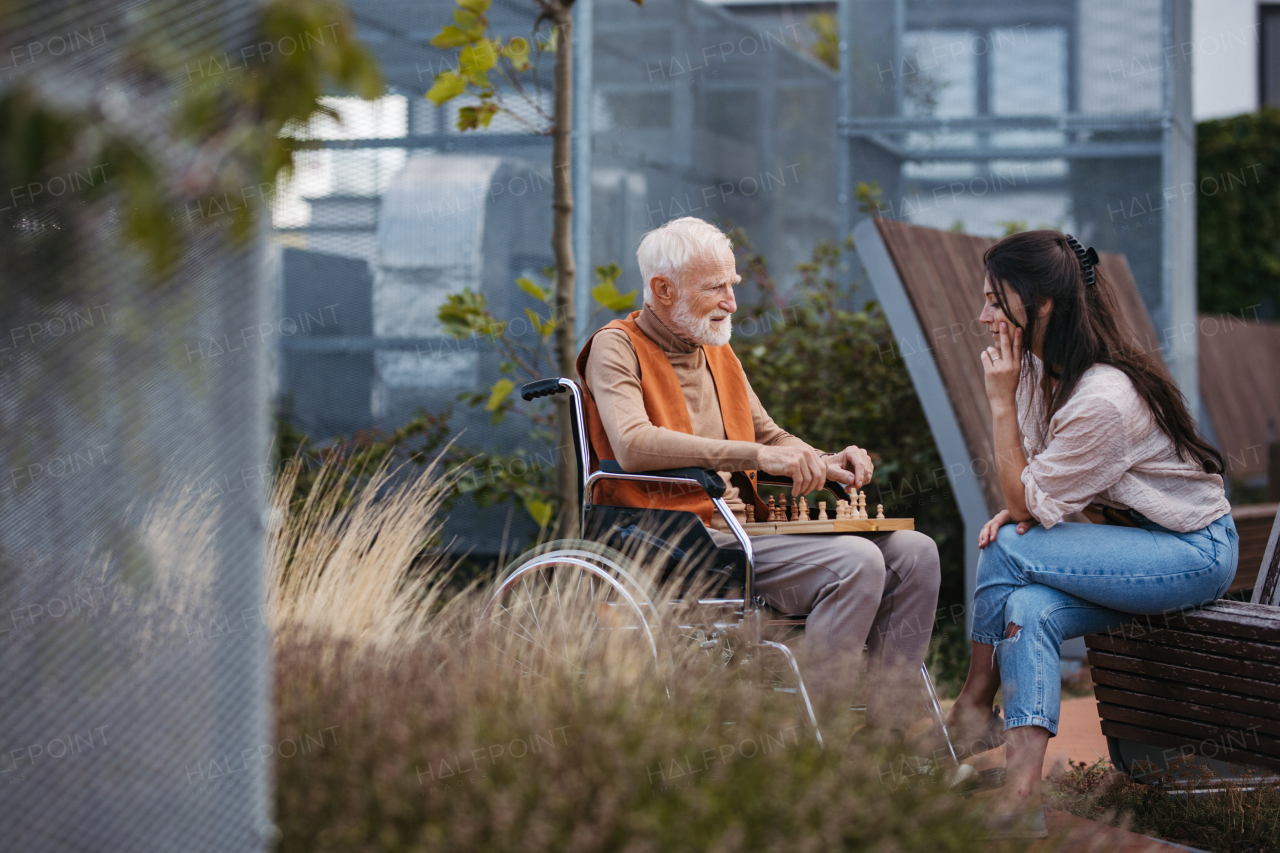 Senior man playing chess outdoors with his daughter. Nursing home client in wheelchair spending quality time with caregiver, enjoying in a game of chess and conversation.