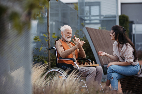 Senior man playing chess outdoors with his daughter. Nursing home client in wheelchair spending quality time with caregiver, enjoying in a game of chess and conversation.