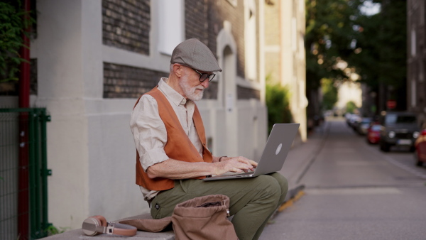 Senior man sitting on street curb working on his laptop outdoors. Video of elderly man using digital technologies, working on a notebook. Concept of seniors and digital skills.