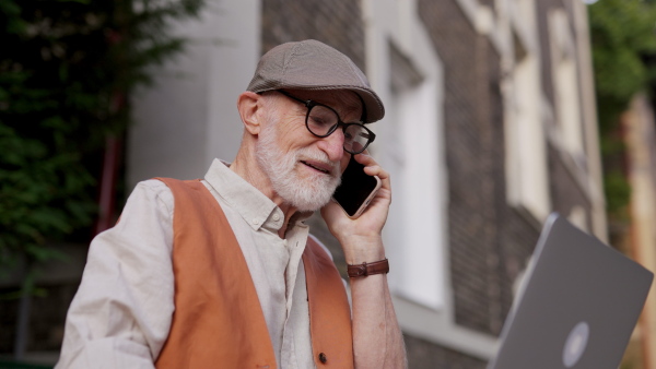 Senior man sitting on street curb working on his laptop outdoors. Video of elderly man using digital technologies, working on a notebook. Concept of seniors and digital skills.