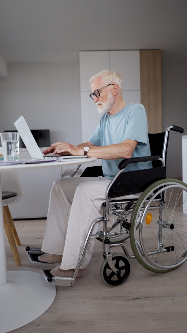 Senior man in a wheelchair working from home during retirement. Portrait of elderly man using digital technologies, working on a laptop. Concept of seniors and digital skills.