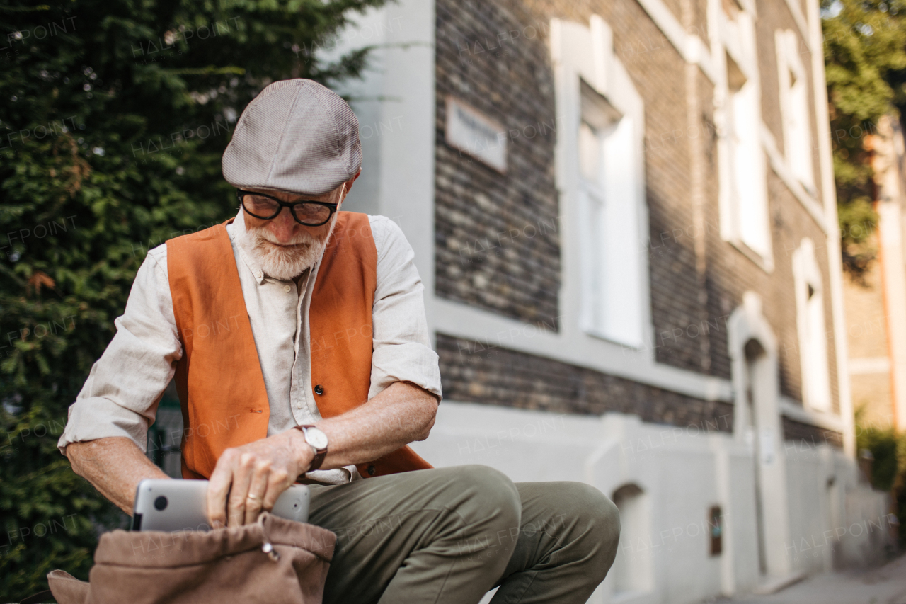 Senior man sitting on street curb in the city. Portrait of elderly man searching for things in a backpack.