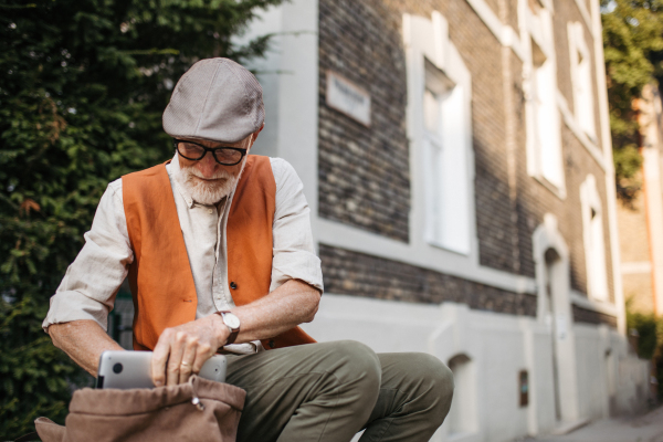 Senior man sitting on street curb in the city. Portrait of elderly man searching for things in a backpack.