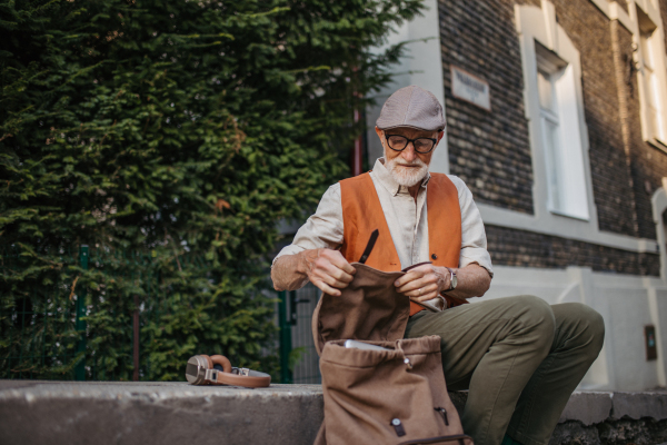 Senior man sitting on street curb working on his laptop outdoors. Portrait of elderly man using digital technologies, working with notebook. Concept of seniors and digital skills.