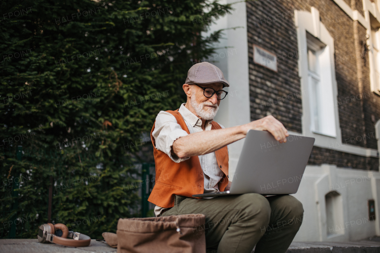 Senior man sitting on street curb working on his laptop outdoors. Portrait of elderly man using digital technologies, working with notebook. Concept of seniors and digital skills.