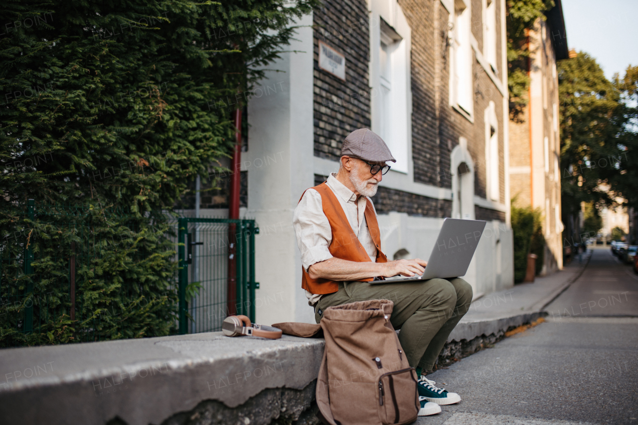 Senior man sitting on street curb working on his laptop outdoors. Portrait of elderly man using digital technologies, working on a notebook. Concept of seniors and digital skills.