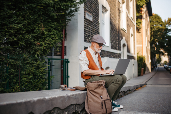 Senior man sitting on street curb working on his laptop outdoors. Portrait of elderly man using digital technologies, working on a notebook. Concept of seniors and digital skills.