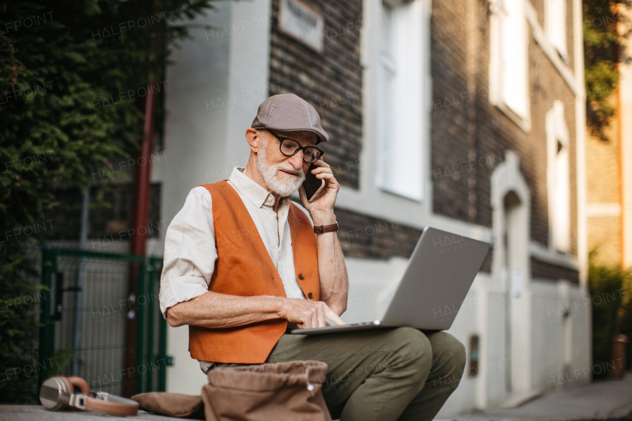 Senior man sitting on street curb working on his laptop outdoors. Portrait of elderly man using digital technologies, working on a notebook. Concept of seniors and digital skills.