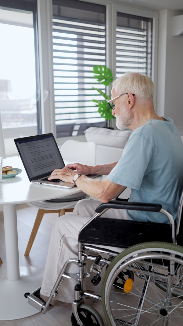 Senior man in a wheelchair working from home during retirement. Portrait of elderly man using digital technologies, working on a laptop. Concept of seniors and digital skills.