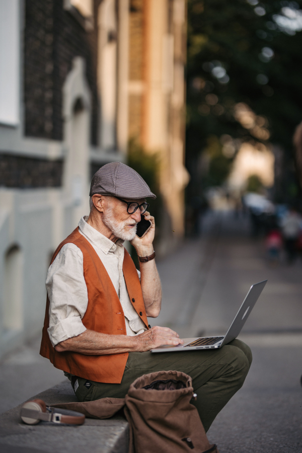 Senior man sitting on street curb working on his laptop outdoors. Portrait of elderly man using digital technologies, working with notebook and smartphone. Concept of seniors and digital skills.