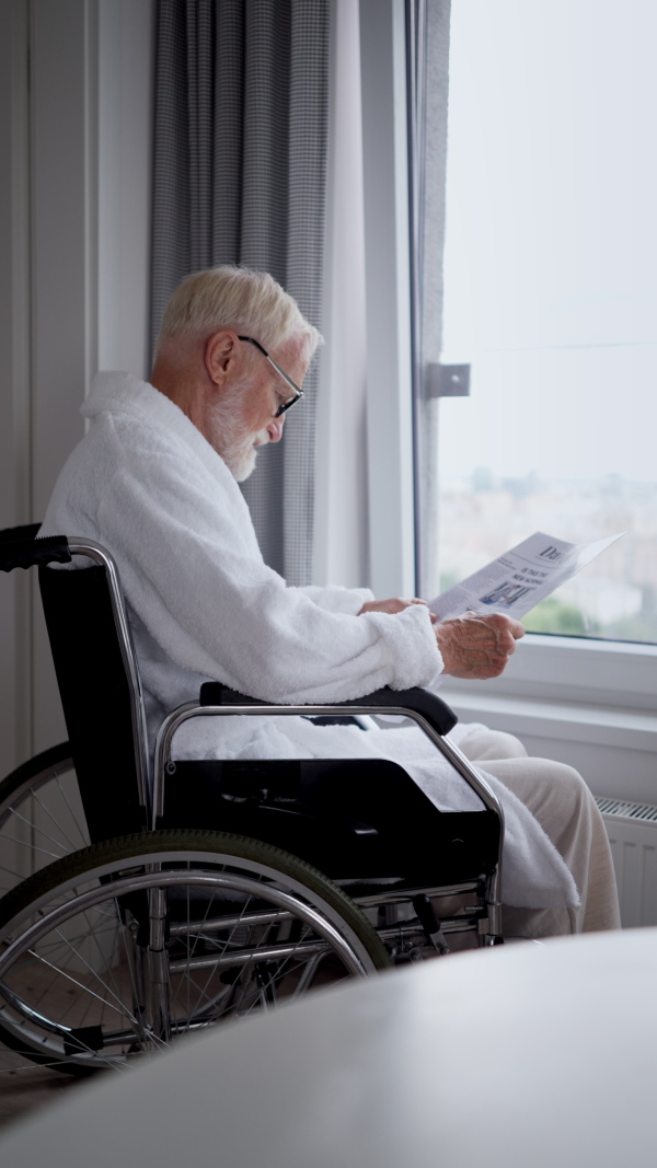 Elderly man in a wheelchair reading the newspaper in his robe in the morning. Concept of loneliness and dependence of retired people.