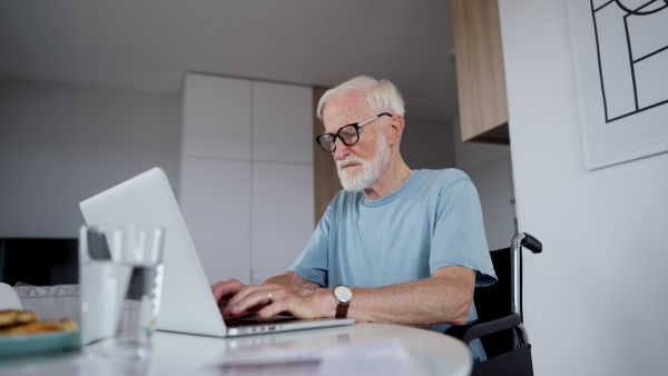 Senior man in a wheelchair working from home during retirement. Video of elderly man using digital technologies, working on a laptop. Concept of seniors and digital skills.