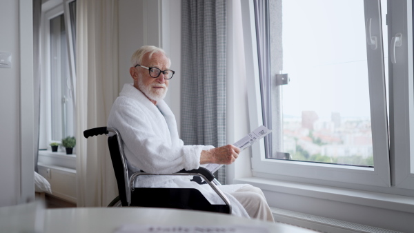 Elderly man in a wheelchair reading the newspaper in his robe in the morning. Concept of loneliness and dependence of retired people.