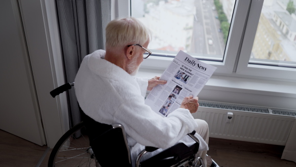 Elderly man in a wheelchair reading the newspaper in his robe in the morning. Concept of loneliness and dependence of retired people.