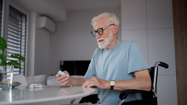 Senior man in wheelchair taking pills at home. Chronically ill man taking medication, reading the label on the bottle.